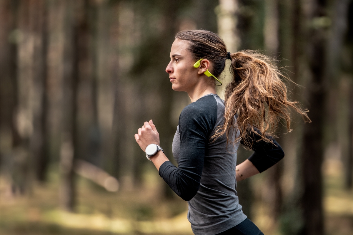 Nainen juoksemassa metsäpolulla, yllään kirkkaankeltaiset Suunto Sonic -luujohdekuulokkeet ja Suunto Race S -älykello.